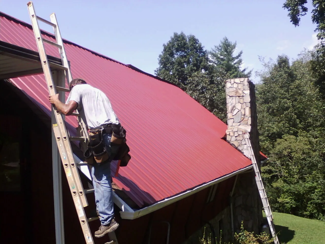 Roofer on a ladder beside a red metal roof and stone chimney — Soffit & Fascia Repair by Roofing Brothers in Varna